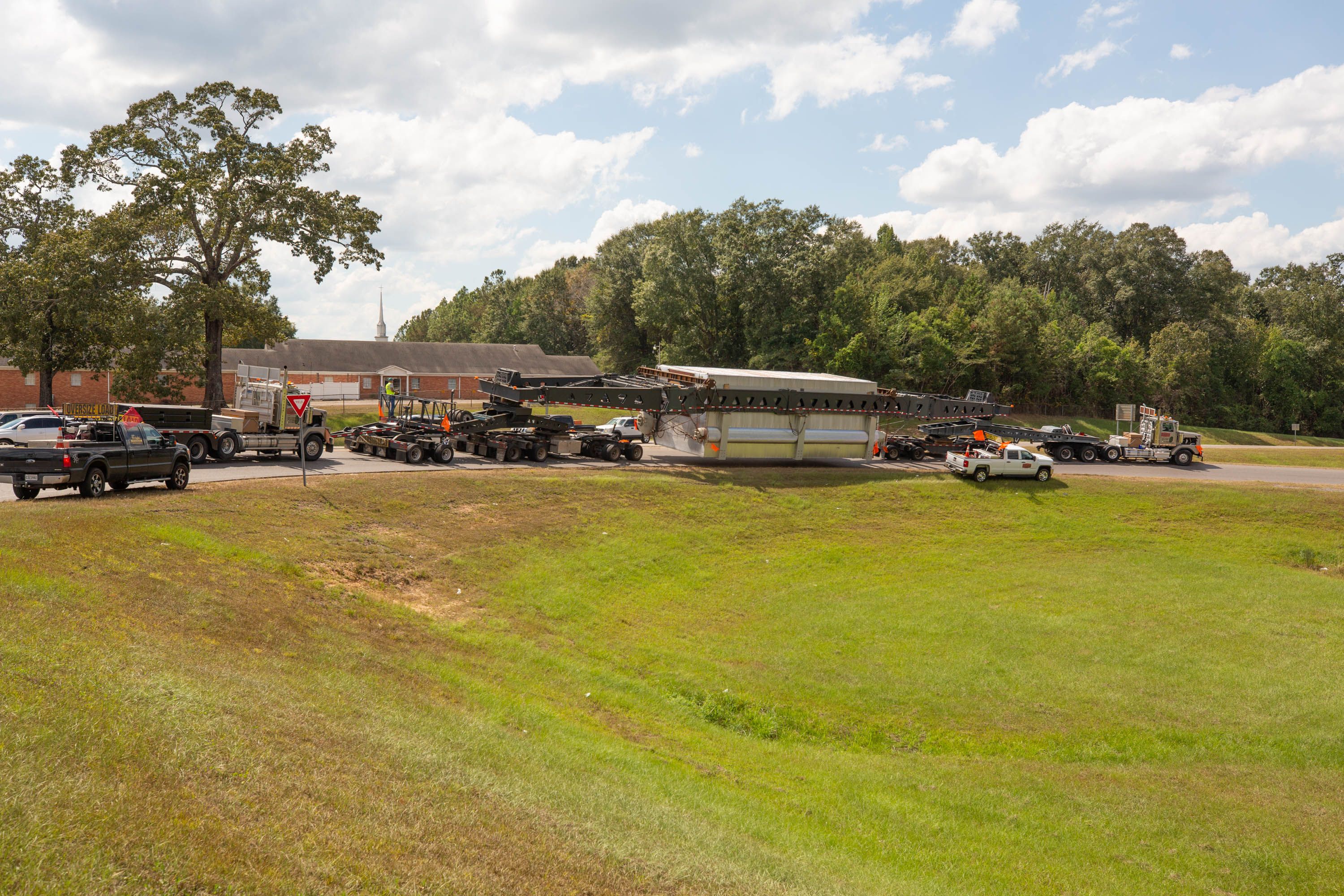 Row of oversize trucks and pilot cars parked alongside grassy bend